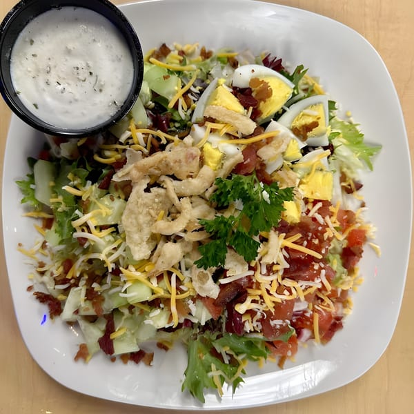 A large salad with lettuce, cheese, bacon, hard-boiled eggs, and fried seafood, with a side of ranch dressing.