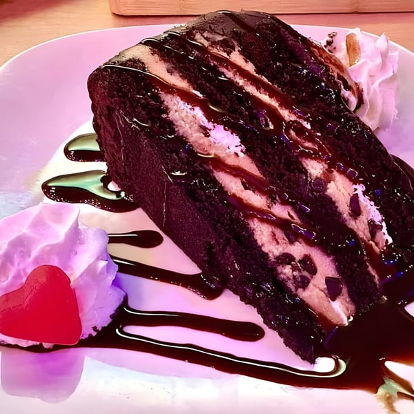 A slice of chocolate cake with cream filling, chocolate chips, and chocolate drizzle, served with whipped cream and a red heart candy.