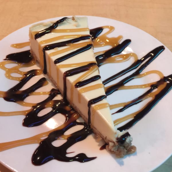 A slice of cheesecake drizzled with chocolate and caramel sauce on a white plate.
