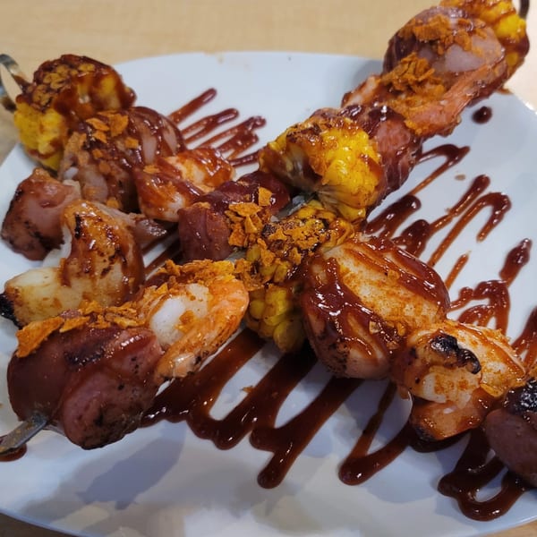 Two skewers of shrimp, sausage, and corn on the cob, drizzled with barbecue sauce.