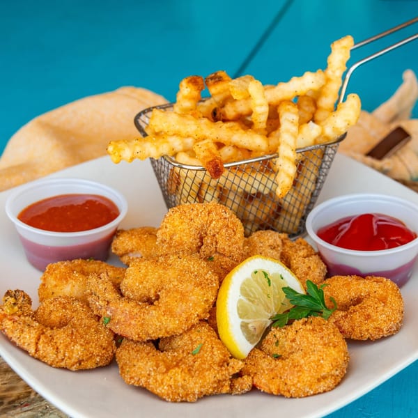 Crispy fried shrimp, crinkle-cut fries in a metal basket, lemon, and two dipping sauces on a white plate.