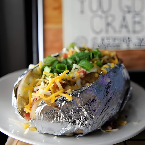 A loaded baked potato, wrapped in foil, topped with cheese, bacon, sour cream, and green onions.