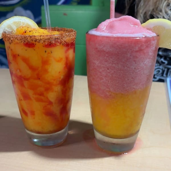 Two colorful frozen drinks, one orange with a chili-lime rim, the other pink and orange, both with lemon slices.